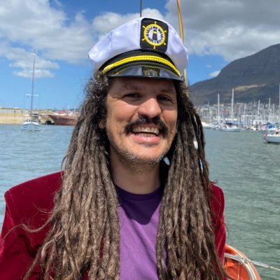 Man with brown moustache and dreadlocks wearing a captain's hat with water and moored sailboats in the background